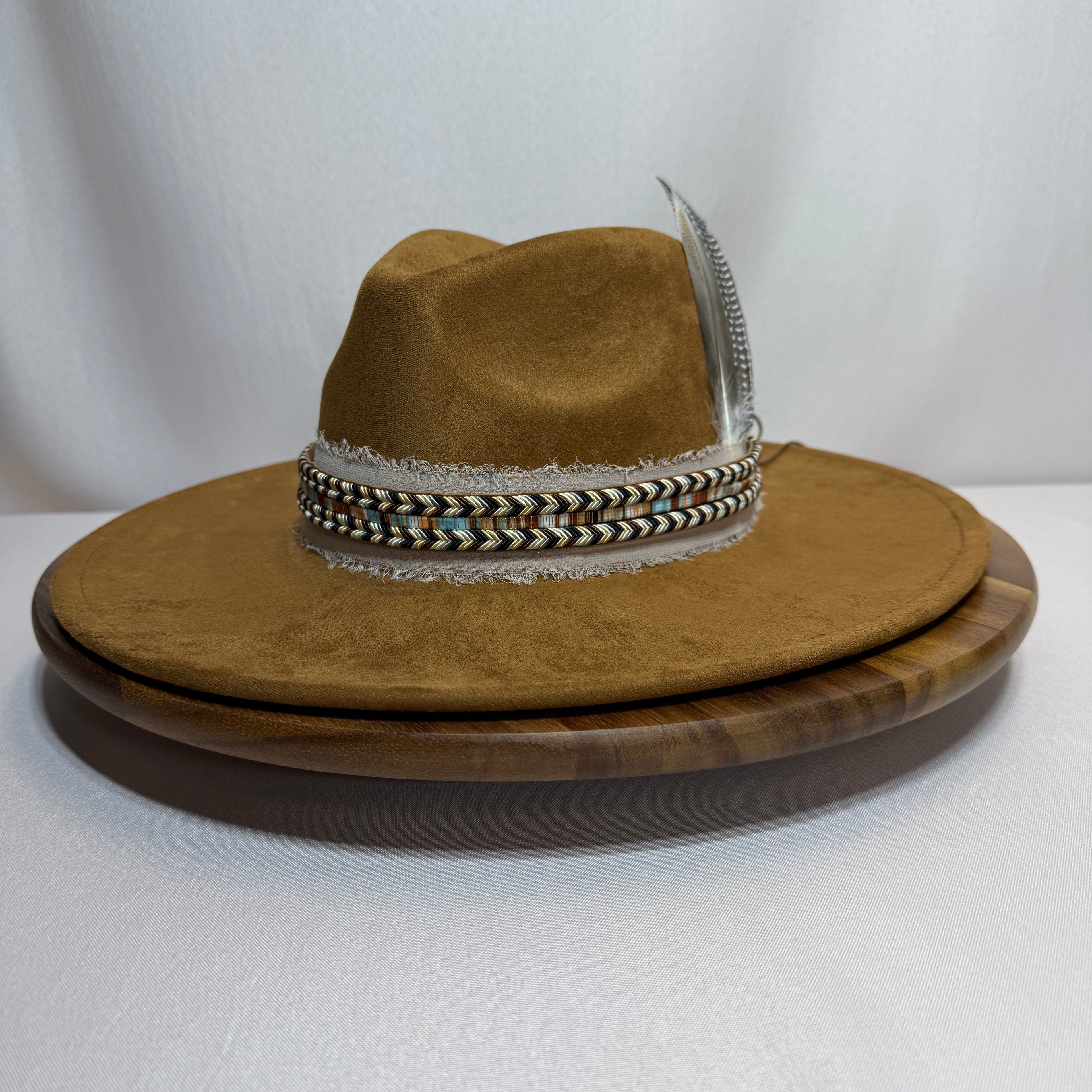 A brown felt cowboy hat with a braided multicolor band and a silver feather, displayed on a wooden turntable against a white fabric backdrop.