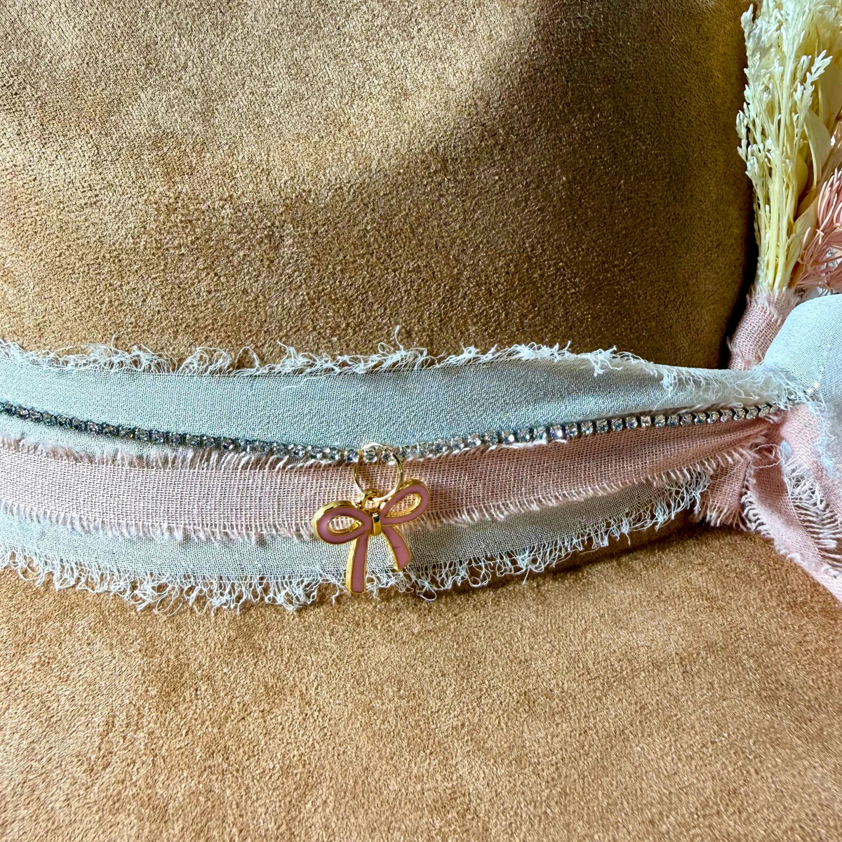 Pink enamel bow charm styled on a camel western hat with soft pink and pale blue ribbon and dried florals