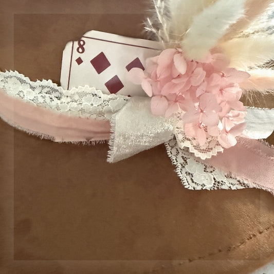 Beautified close-up of the exact distressed eight of diamonds card add-on styled behind pink ribbon, lace and florals on a brown western hat